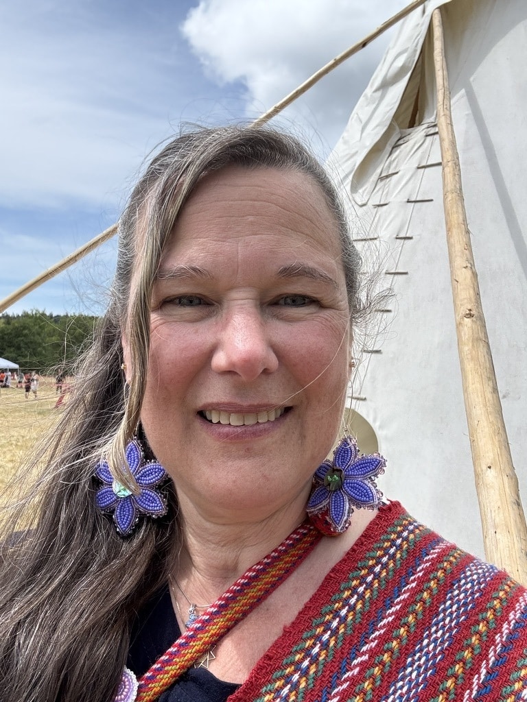 Smiling woman with long gray hair wearing purple flower earrings and a colorful woven shawl, standing beside a white canvas tent outdoors.